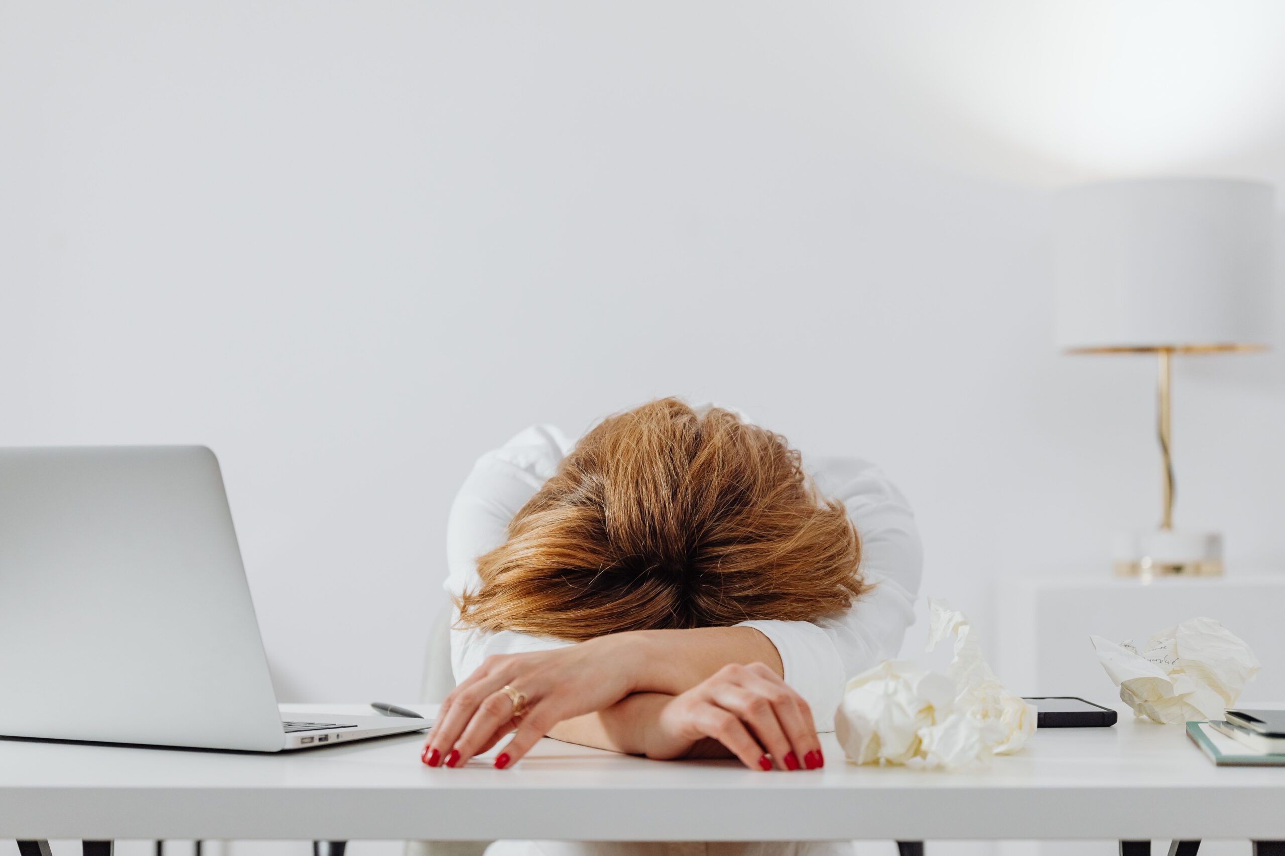 Tired woman resting head on arms at desk with laptop, showing fatigue in modern office.